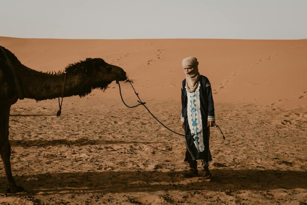 A man in traditional attire leads a camel in the Sahara Desert, Morocco.